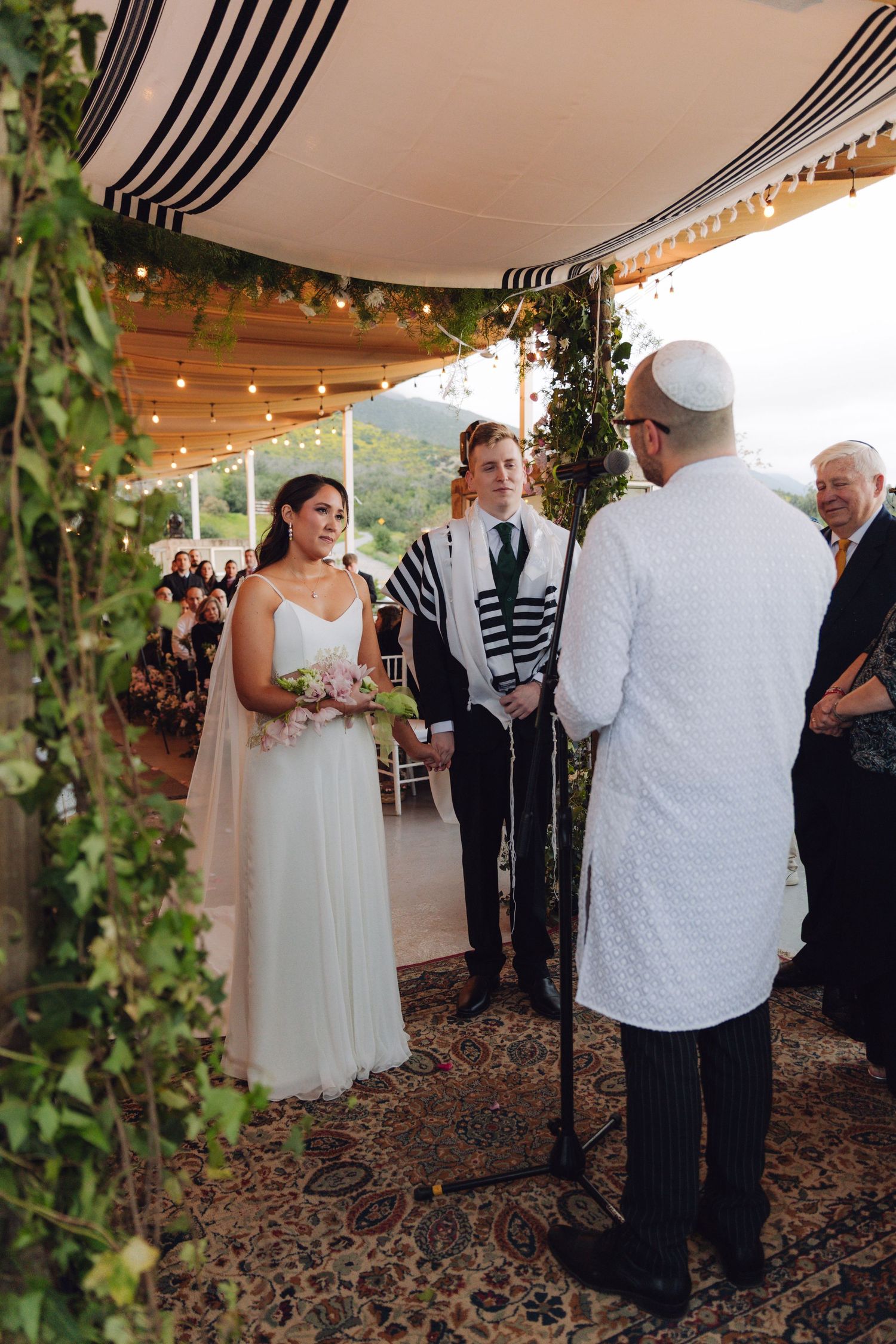 Rabbi speaking to couple under decorated chuppah at outdoor Jewish wedding.
