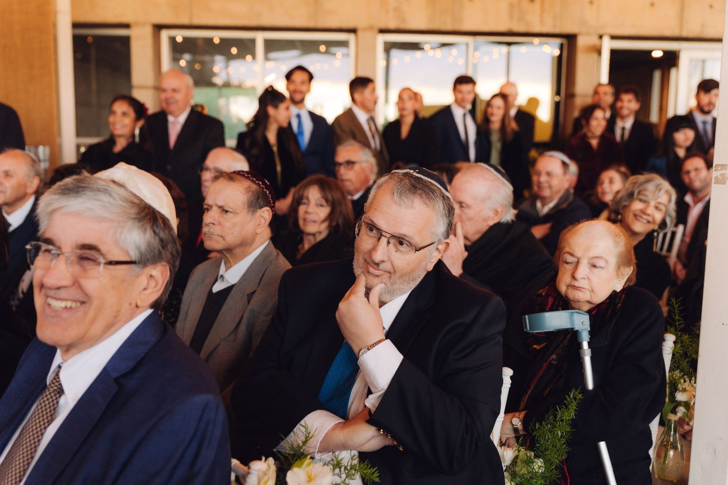 Wedding guests smiling and watching a Jewish ceremony in a warmly lit venue.