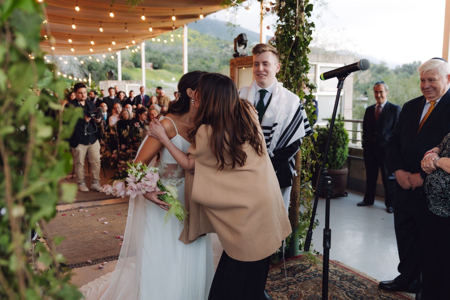 Bride receiving a heartfelt hug from a guest during an outdoor Jewish wedding ceremony.