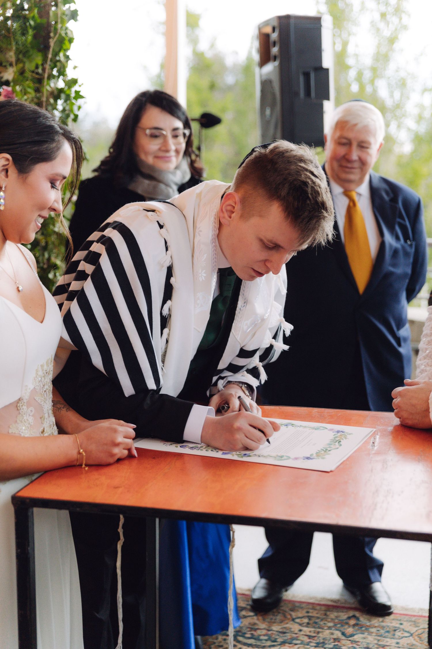 Groom signing the ketubah at a Jewish wedding ceremony with bride and witnesses nearby.