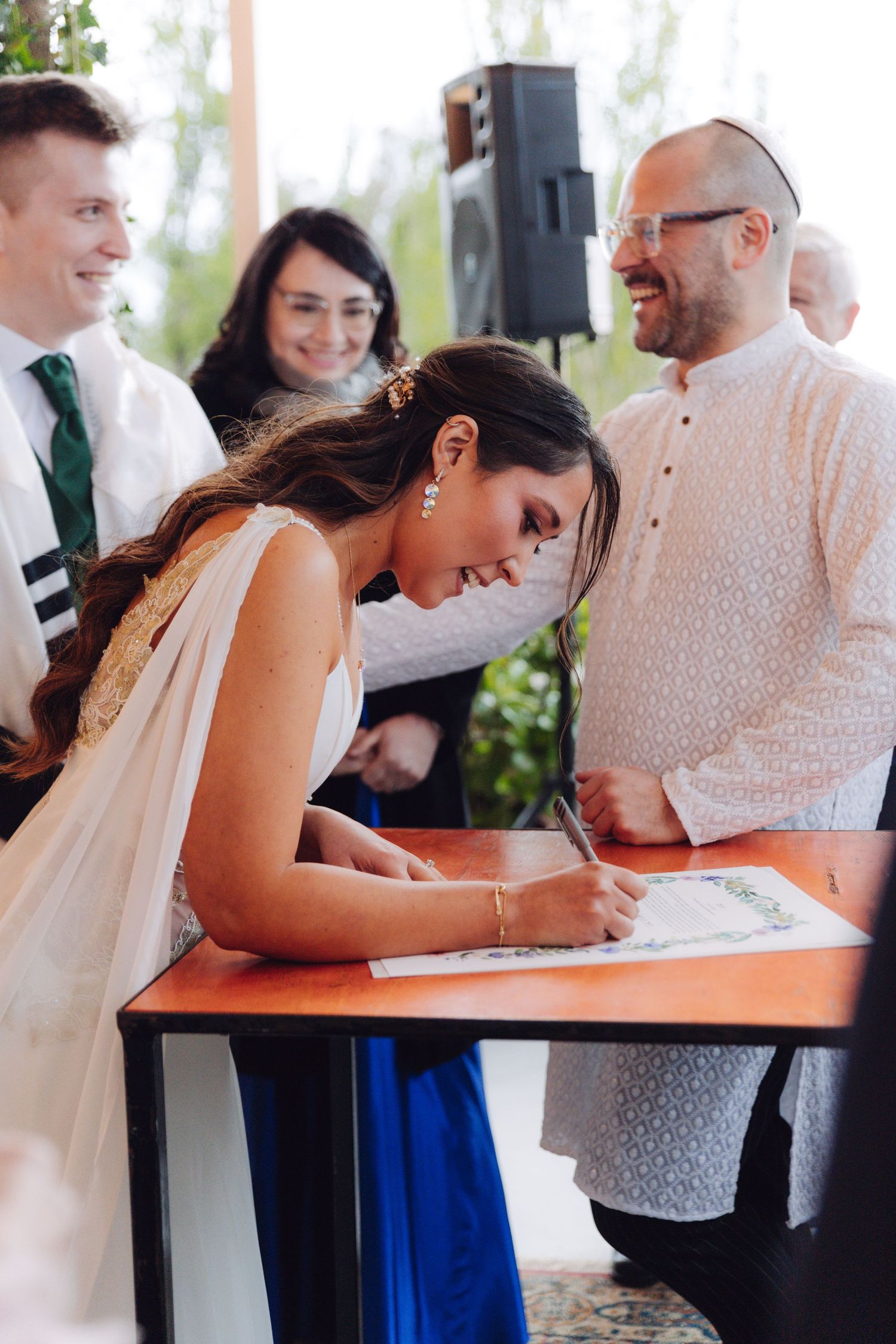 Bride signing the ketubah at a Jewish wedding ceremony while the officiant smiles warmly.