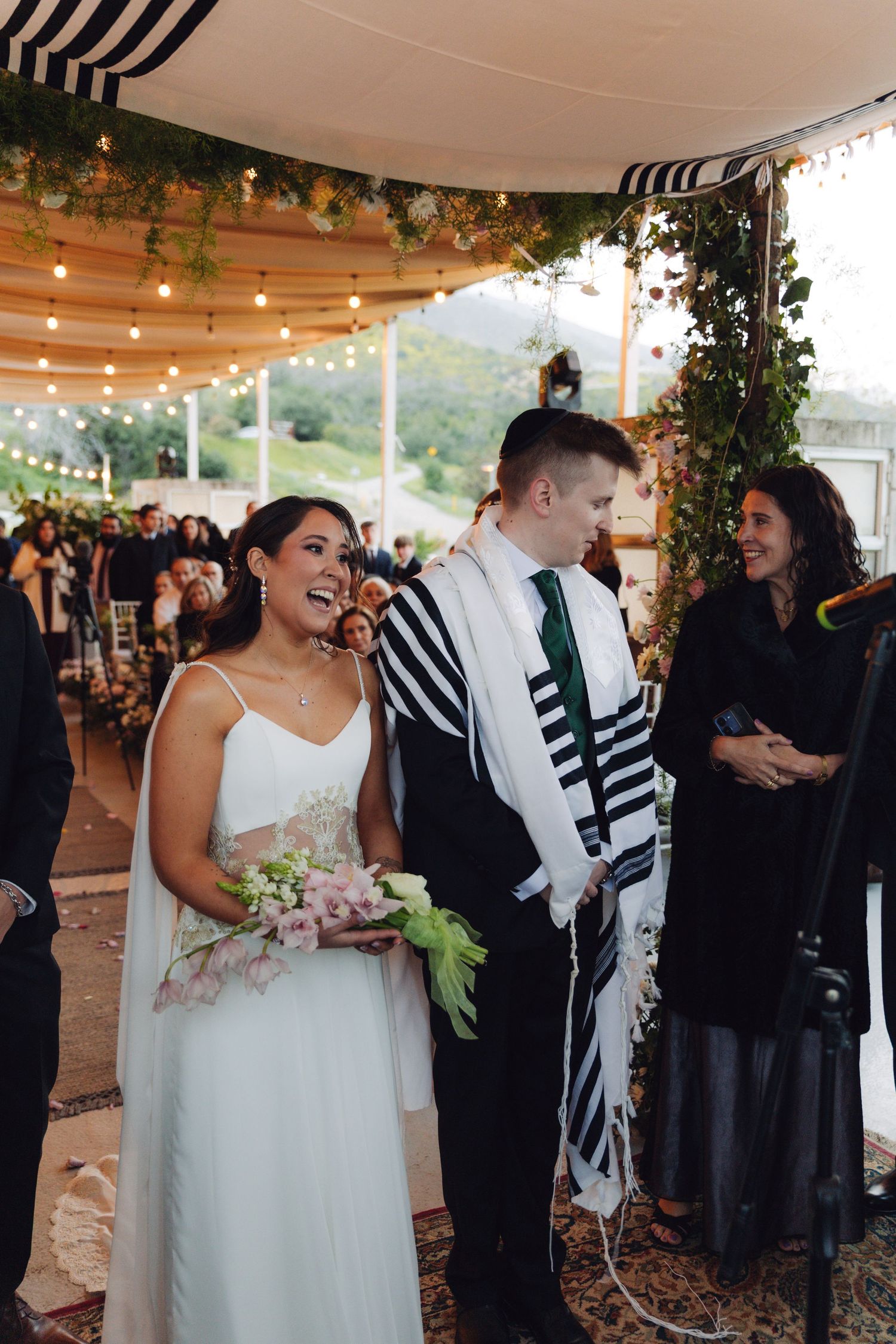 Bride and groom smiling joyfully under a floral chuppah at a Jewish wedding ceremony.