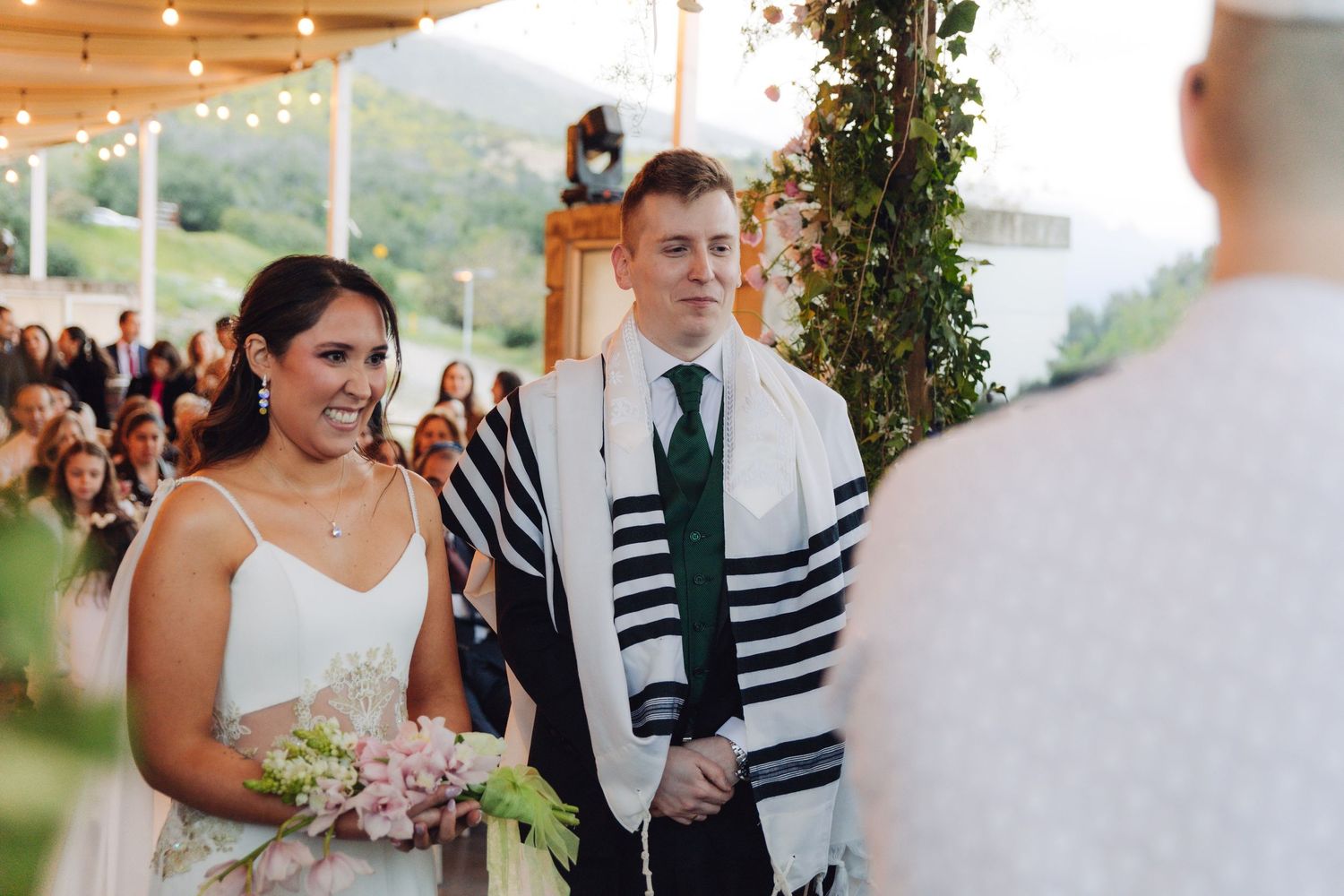 Bride and groom stand together smiling before the officiant at a Jewish wedding ceremony.