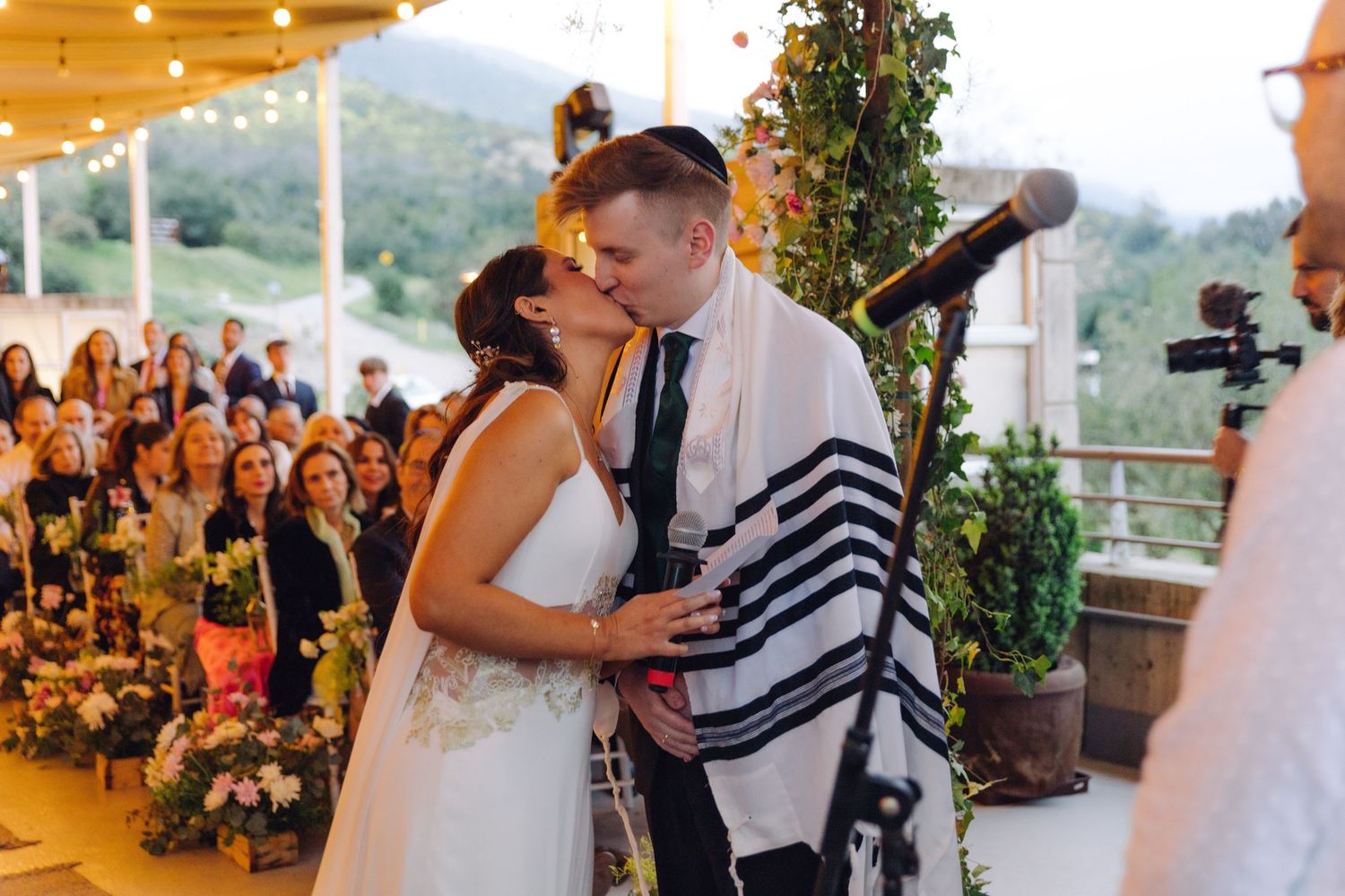 Bride and groom share first kiss under tallit at outdoor Jewish wedding ceremony with guests applauding.