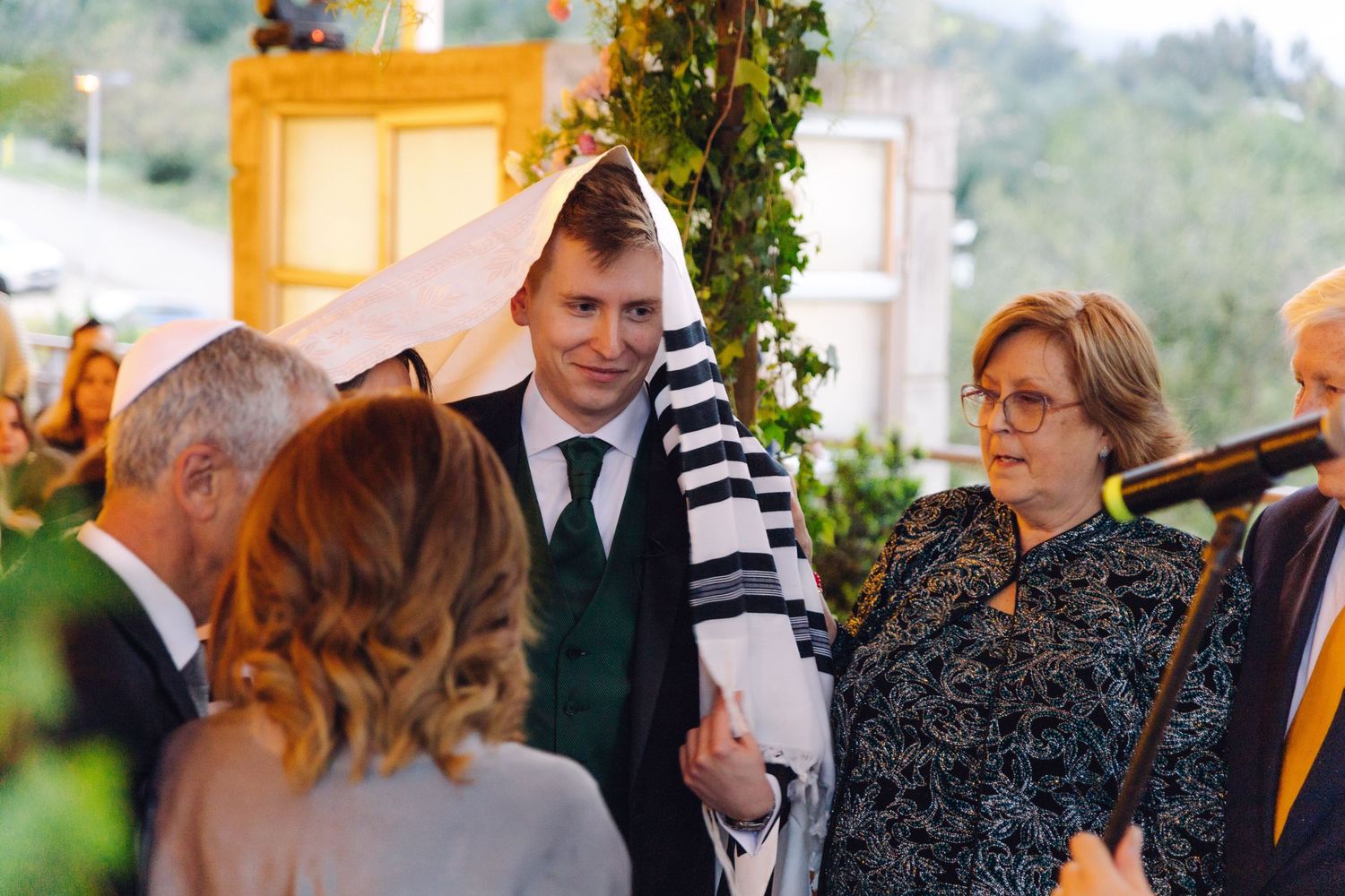 Groom wrapped in tallit greeted by family members during Jewish wedding ceremony outdoors.