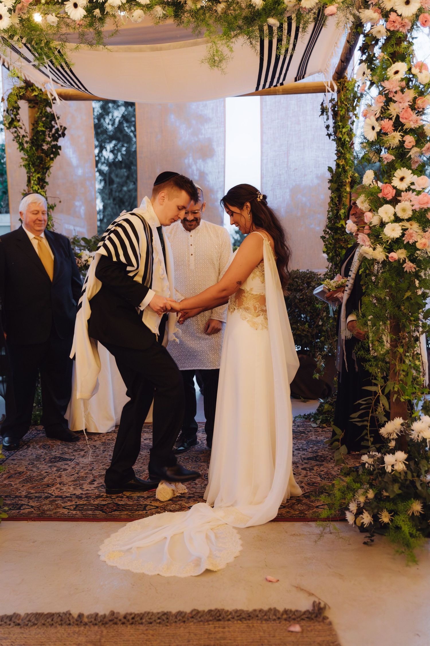 Groom stomps glass under floral chuppah during traditional Jewish wedding ceremony with officiant.