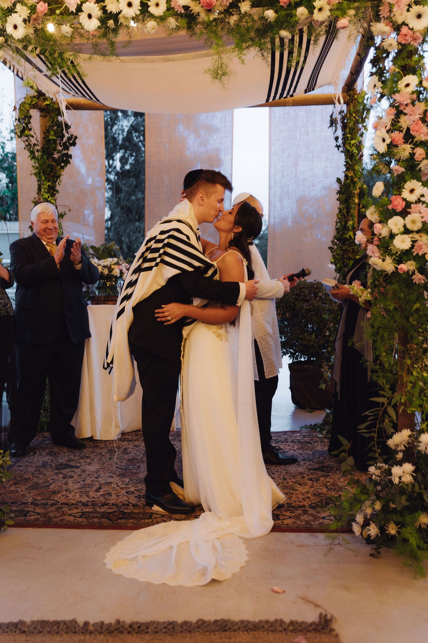 Newlyweds share romantic first kiss under floral chuppah at elegant Jewish wedding ceremony.