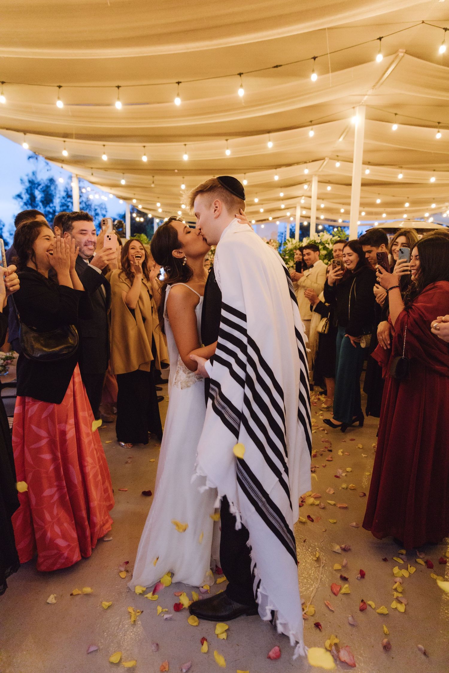 Newlyweds share first kiss at Jewish wedding ceremony as guests celebrate.