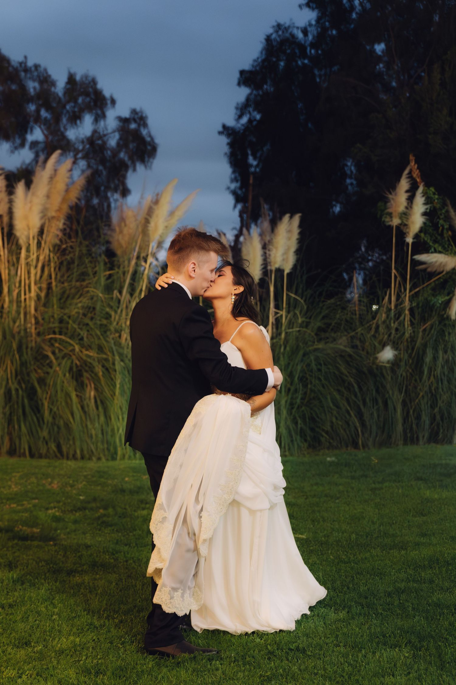 Bride and groom share a passionate kiss on a lush green lawn at dusk with pampas grass behind them.