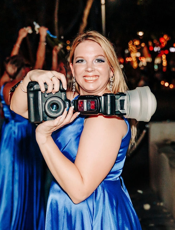 Smiling blonde woman in blue dress holding professional camera with flash at evening event.