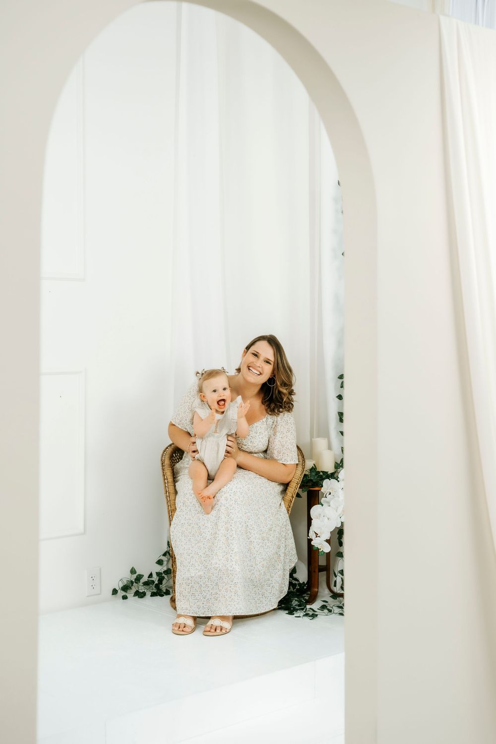 A smiling mother in a white lace dress sits in a wicker chair holding her laughing baby in a bright white room.