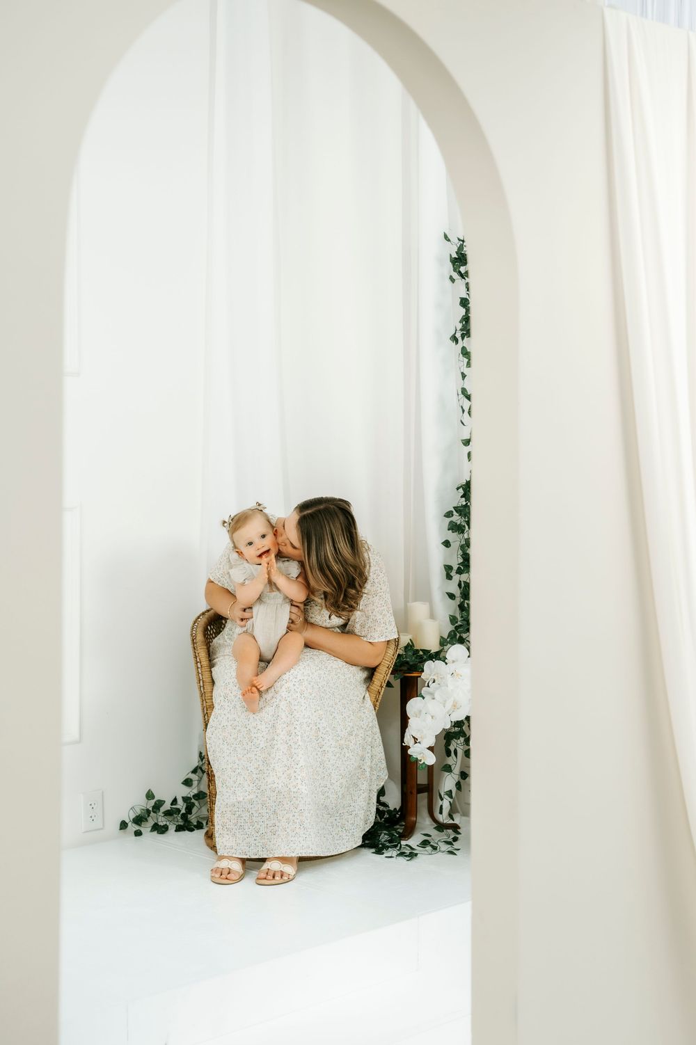 A mother in a white dress kisses her baby girl while seated in a wicker chair under an elegant arch.