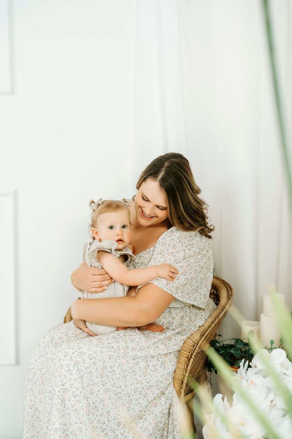 A smiling mother in a floral dress holds her baby in a wicker chair, surrounded by white decor and flowers.