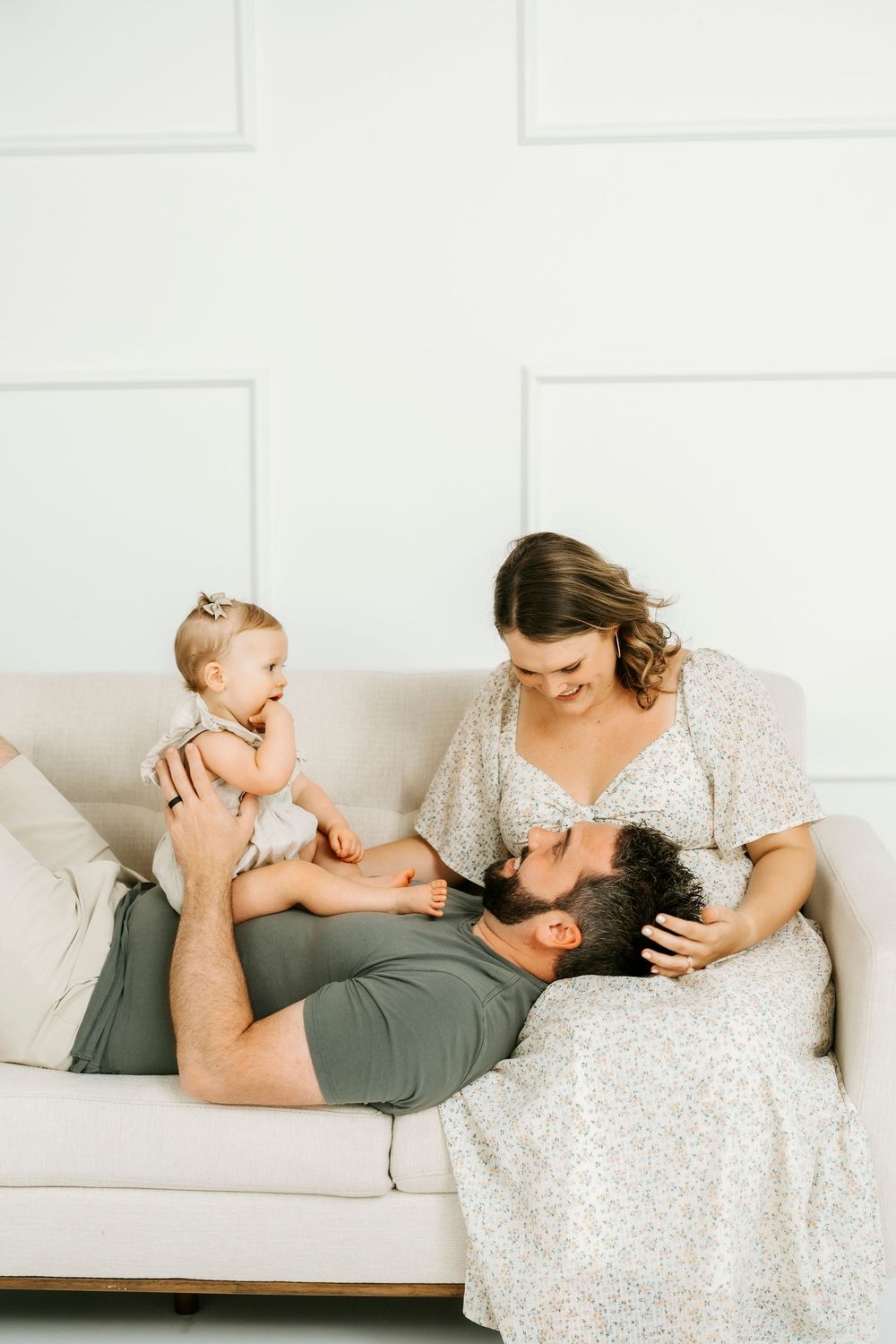 A happy family with a baby girl relaxing on a beige sofa, father lying down while mother smiles warmly in a bright room.