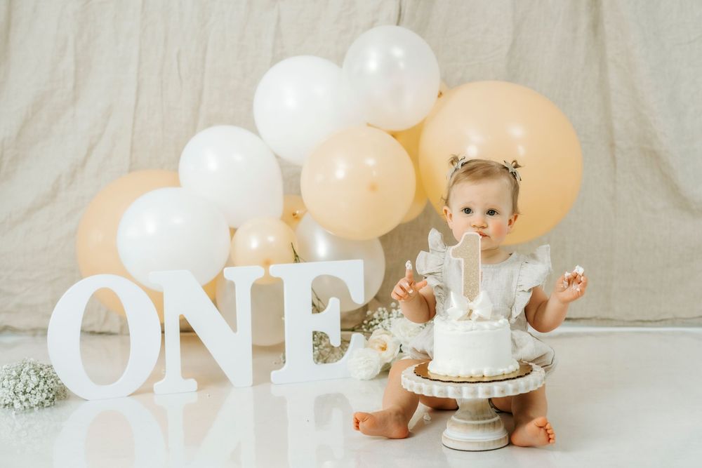 Baby's first birthday cake smash with white and peach balloon garland, ONE sign, and white cake on white floor.