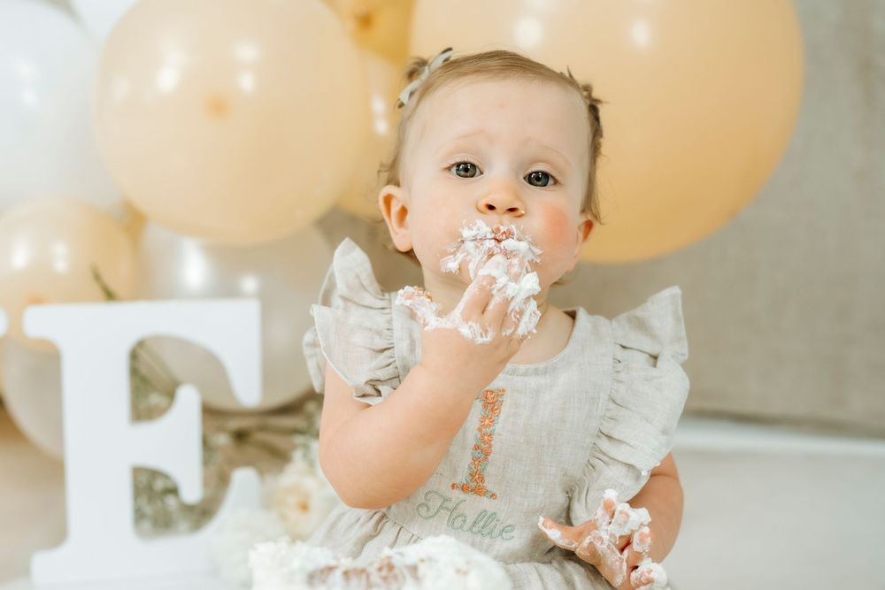 Baby girl in white dress eating smash cake at first birthday party with beige balloons and letter F decoration.