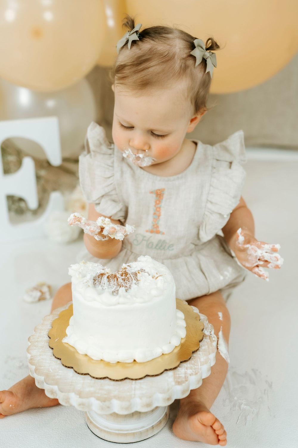 Baby girl in gray ruffle romper smashing her first birthday cake, covered in white frosting with balloons in background.