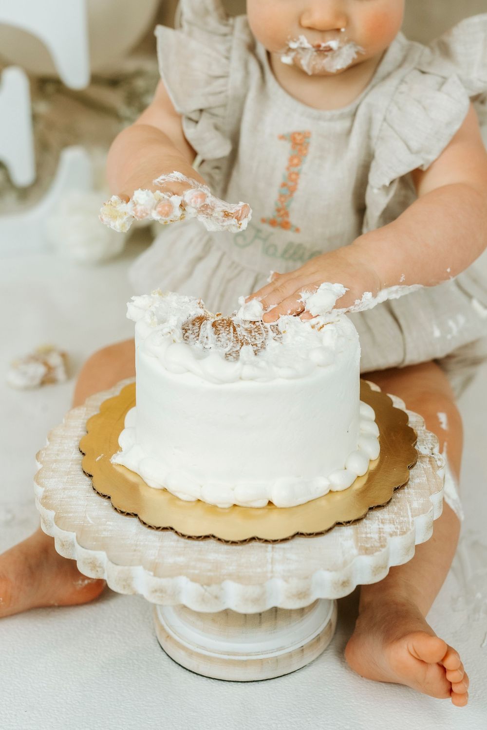 Baby girl in gray dress smashing white first birthday cake, with frosting on hands and face.