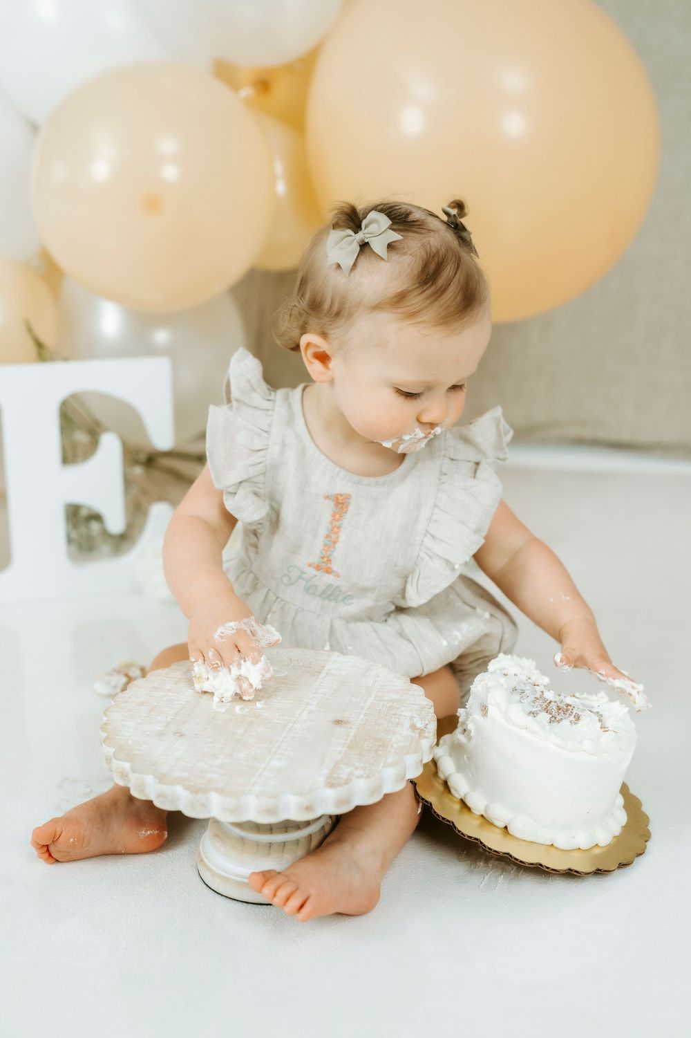 Toddler girl in gray ruffle romper exploring smashed first birthday cake on white cake stand with gold balloons behind.
