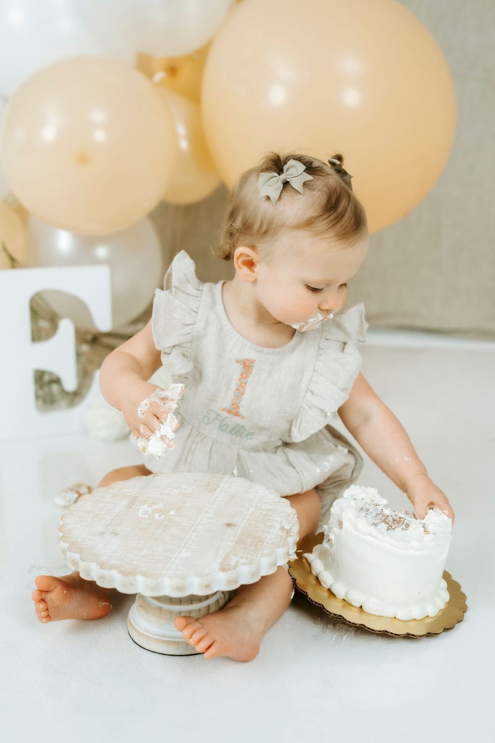 Baby girl in gray romper smashing her first birthday cake surrounded by gold and white balloons.