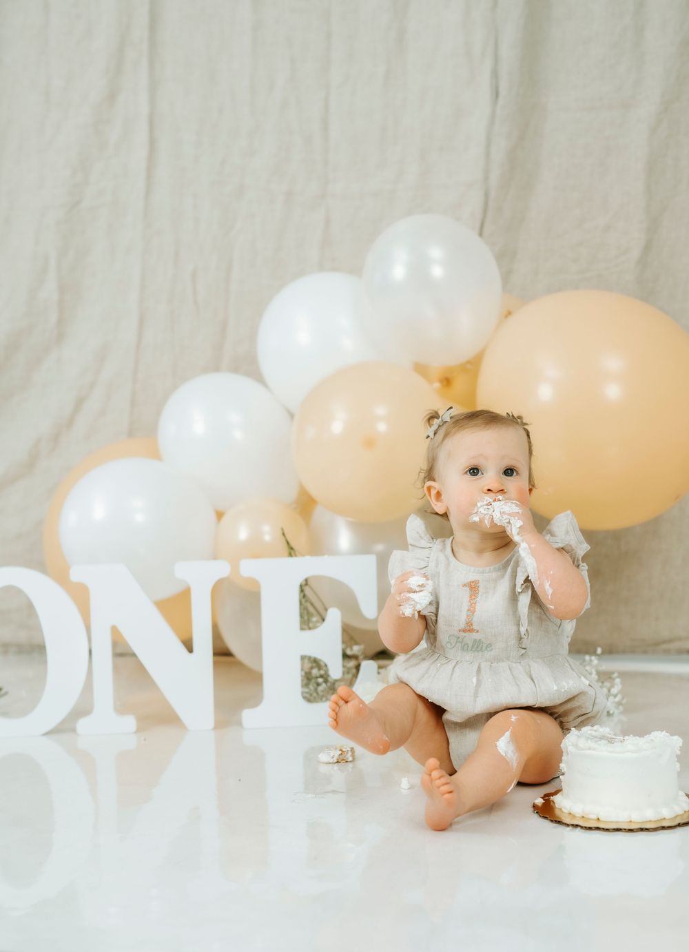 Baby girl celebrating her first birthday with cake smash, surrounded by white and peach balloons and ONE letters.