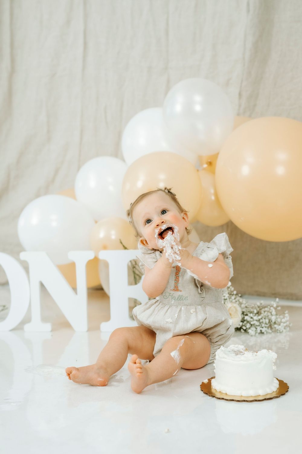 Baby girl smashing her first birthday cake surrounded by white and gold balloons and ONE letters.