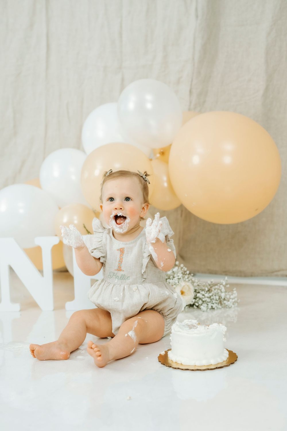 Excited baby girl celebrating first birthday with smash cake and gold and white balloon backdrop.