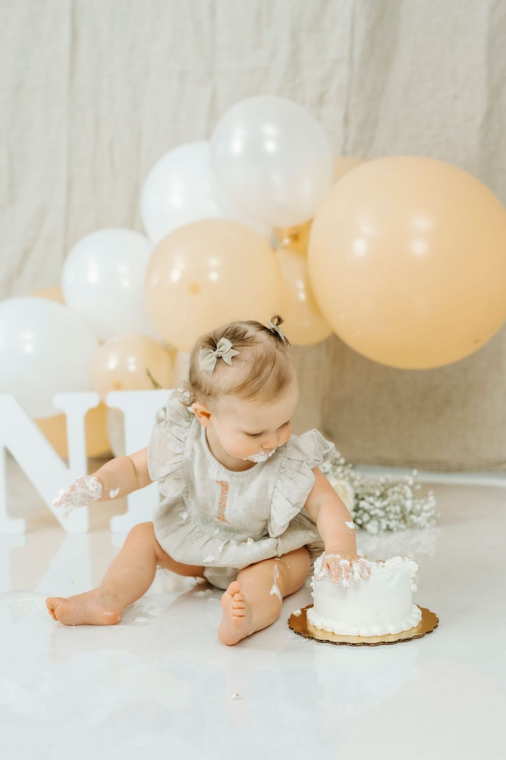Baby girl in gray romper smashing first birthday white cake surrounded by peach and white balloons.