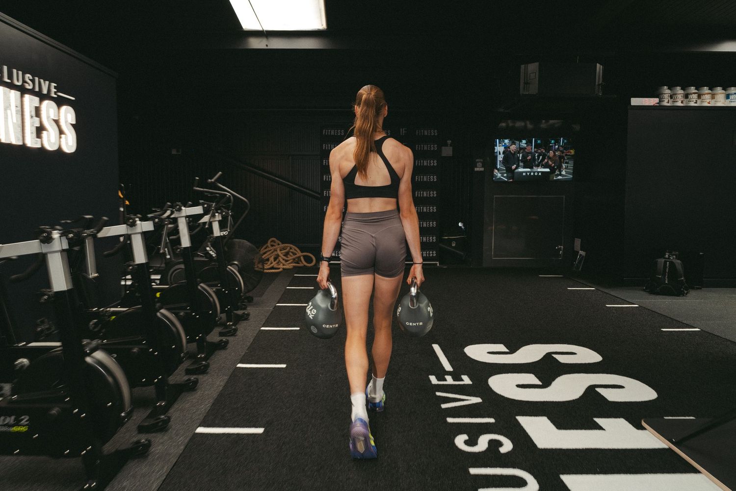 Woman carrying kettlebells walking through a modern gym with fitness equipment and branded flooring.