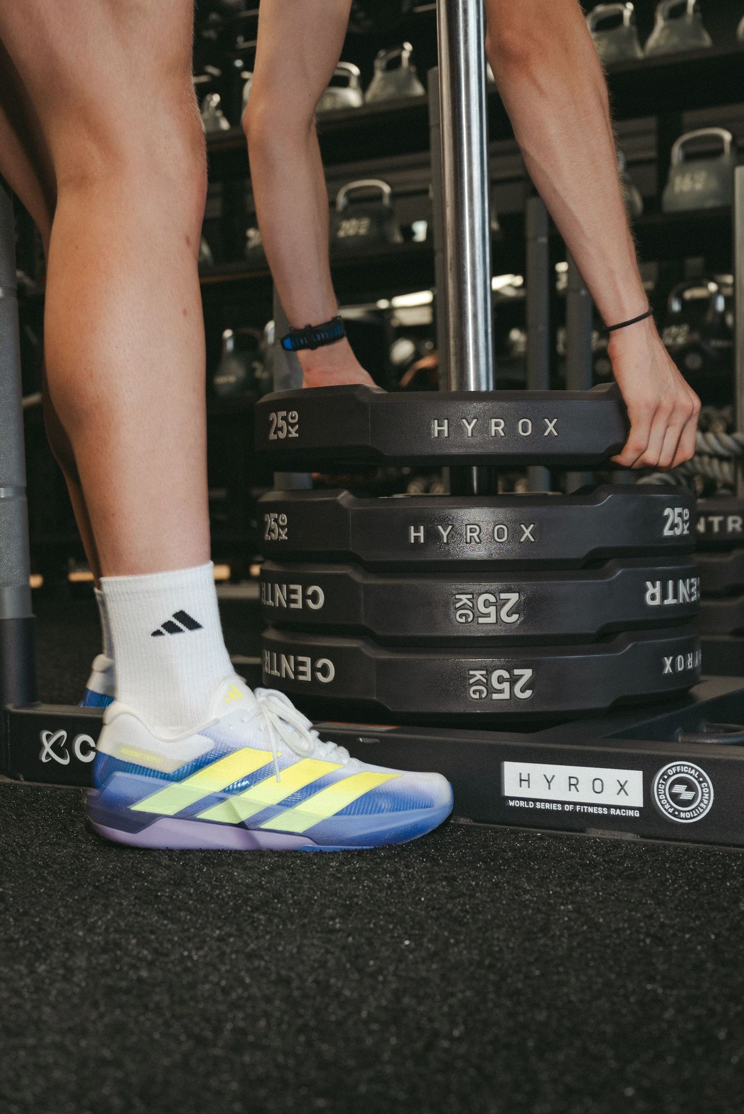 Adidas HYROX training shoes in blue and yellow worn with white socks next to weighted barbell plates in a gym.