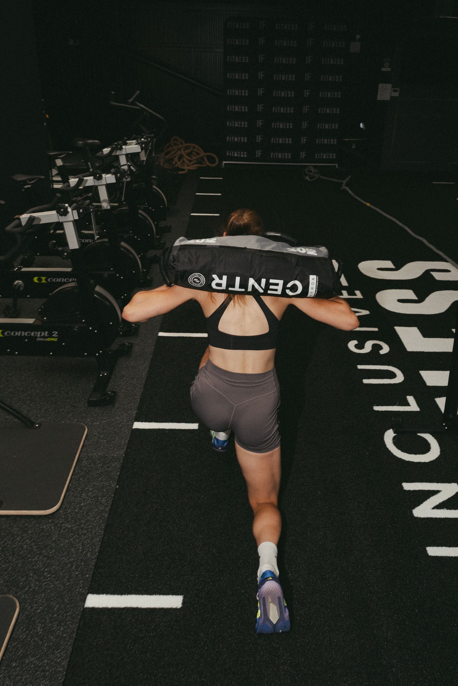 Woman performing lunges with a sandbag on her shoulders in a dark gym with inclusive fitness branding on the floor.
