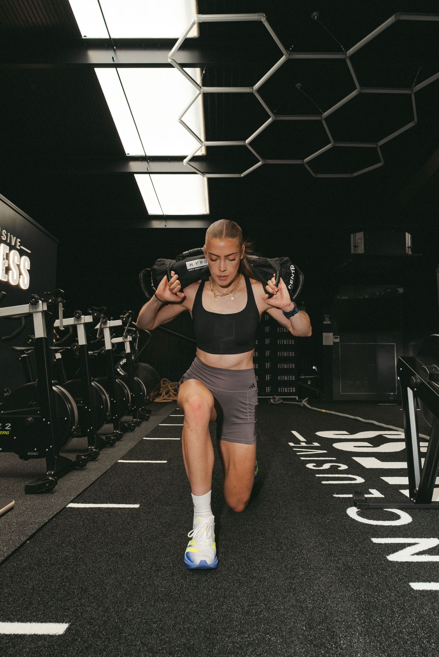 Athletic woman performing lunges with sandbags on shoulders in a modern dark gym wearing Adidas shoes.