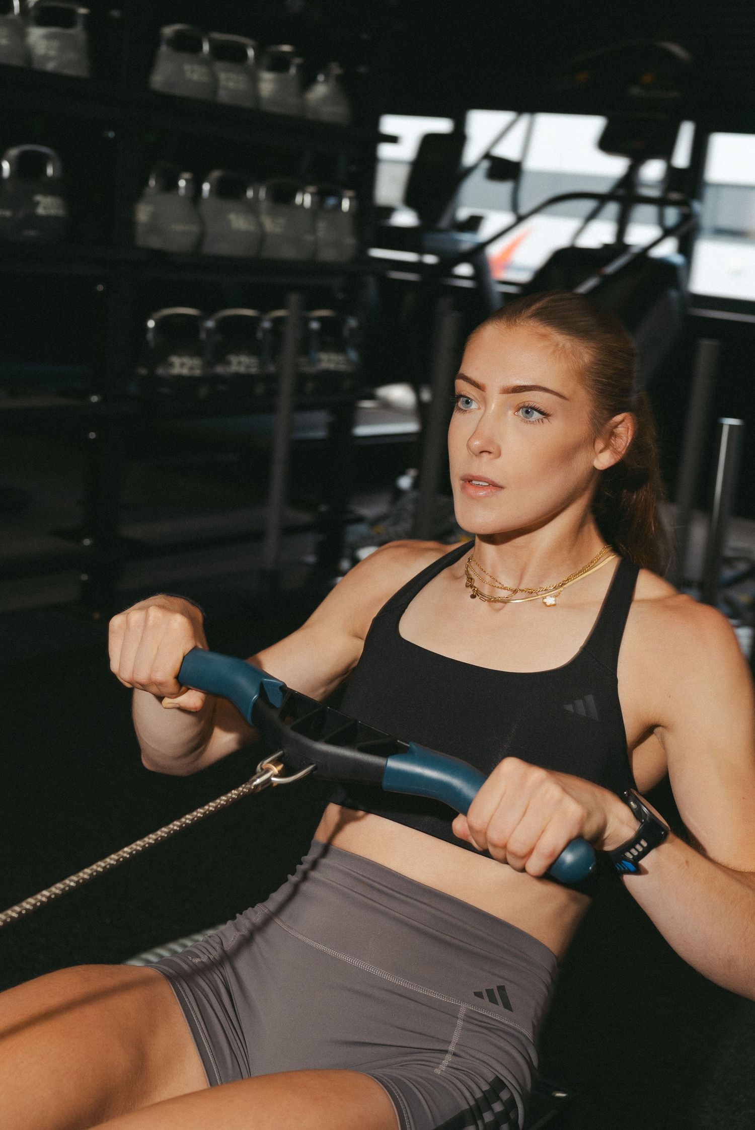 Woman in Adidas sports bra and shorts using a rowing machine in a modern gym.