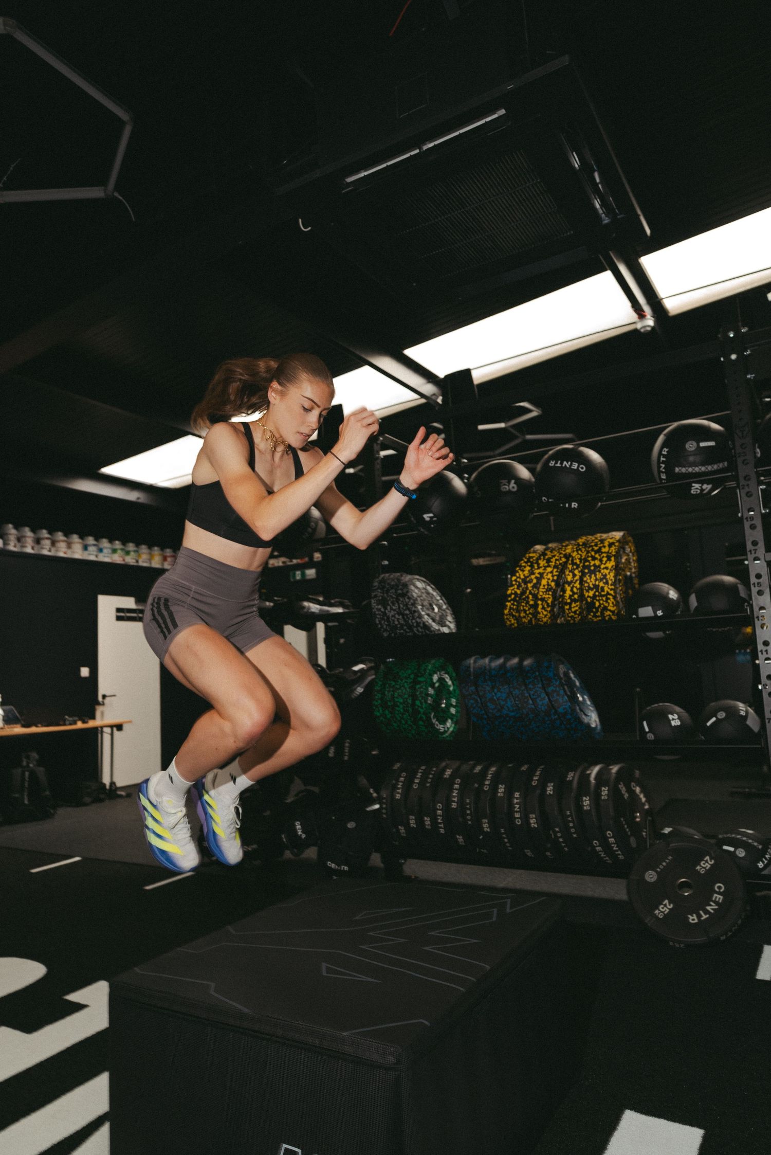 Woman performing a box jump exercise in a dark gym with weight equipment and barbells in the background.