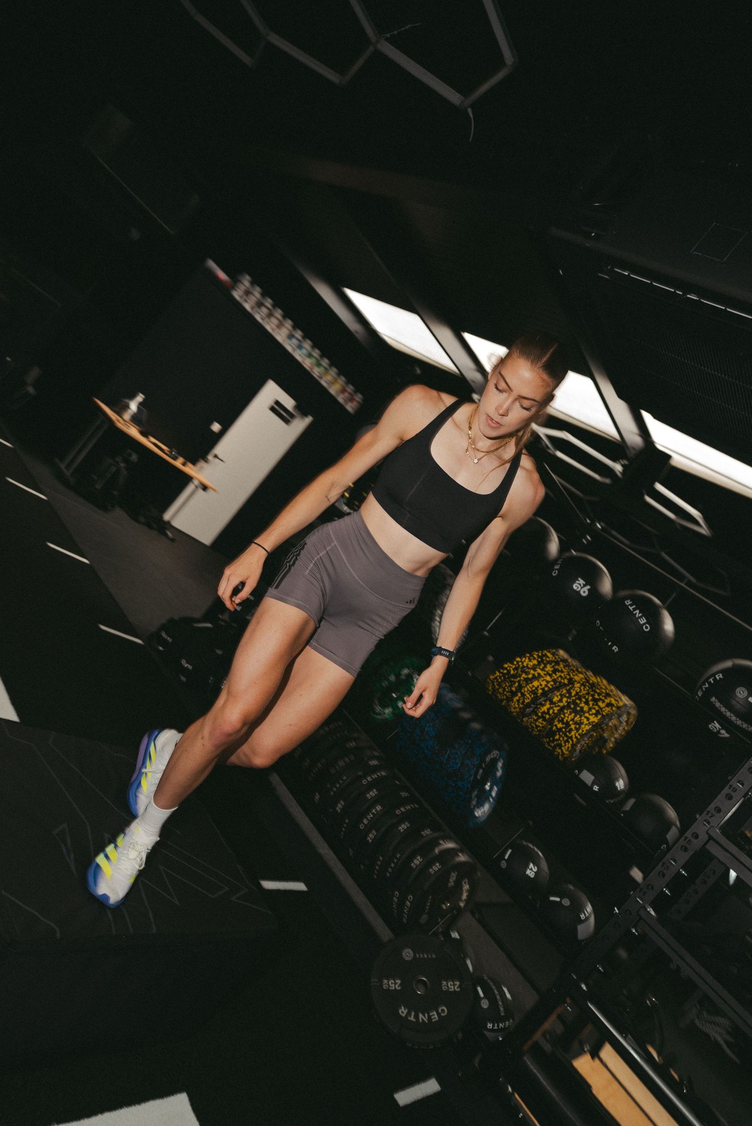 A fit woman in athletic wear stands near a dumbbell rack in a modern dark gym.