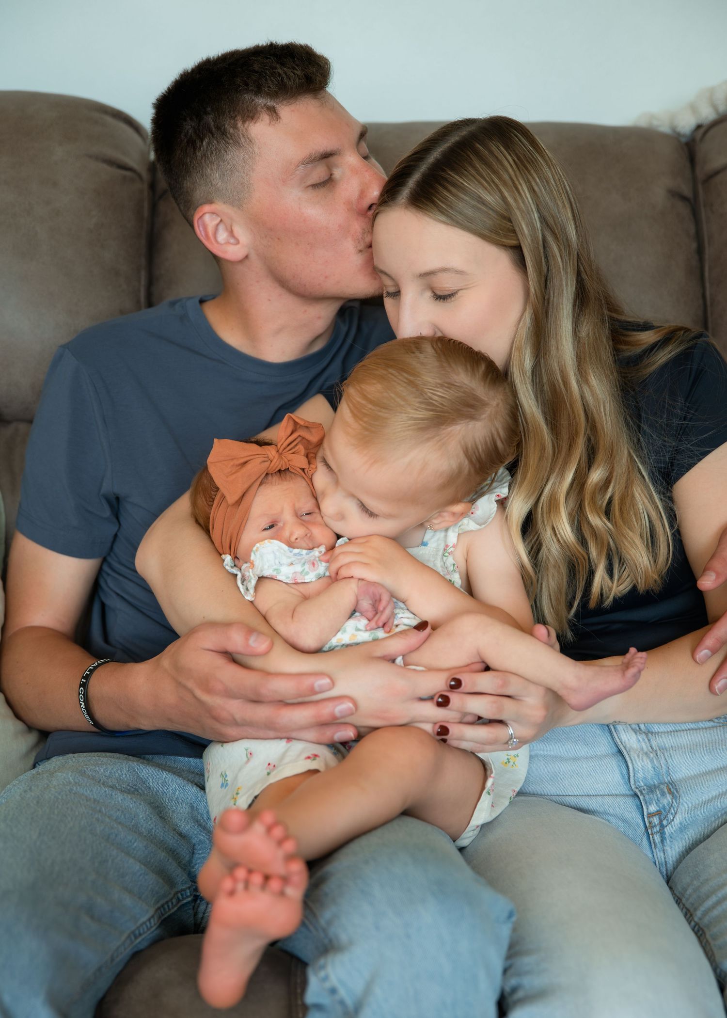 Young couple sitting on couch kissing while holding toddler and newborn baby wrapped in orange bow.