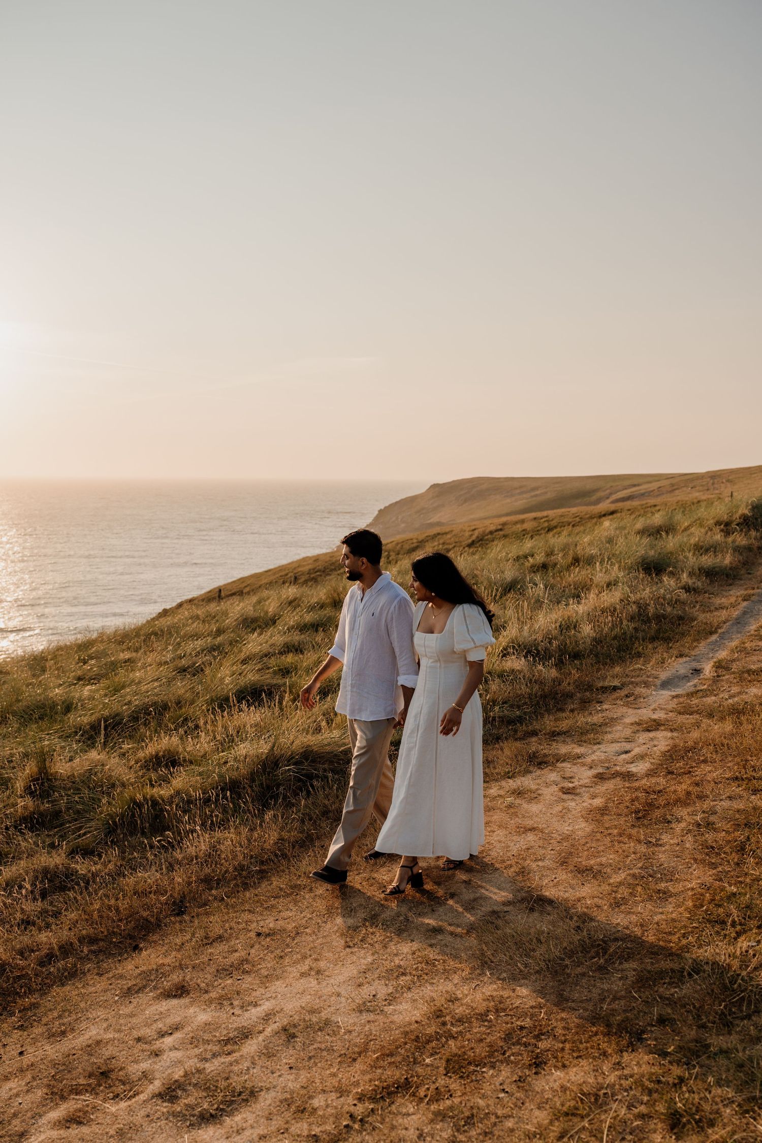 Couple in white outfits walking along the cliffs at Holywell Bay at golden sunset with ocean views.