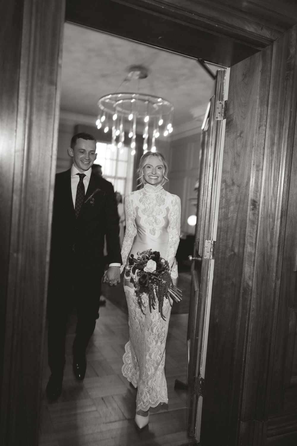 Black and white photo of a smiling bride in lace gown and groom in suit walking through elegant doorway with chandelier.