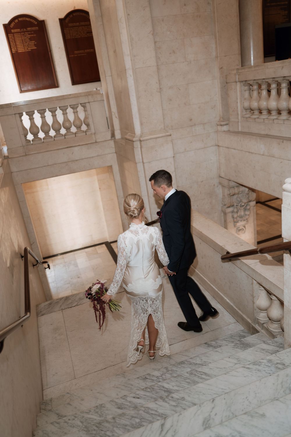 Bride in lace dress and groom in dark suit walk up marble staircase in elegant venue, bride holding bouquet.