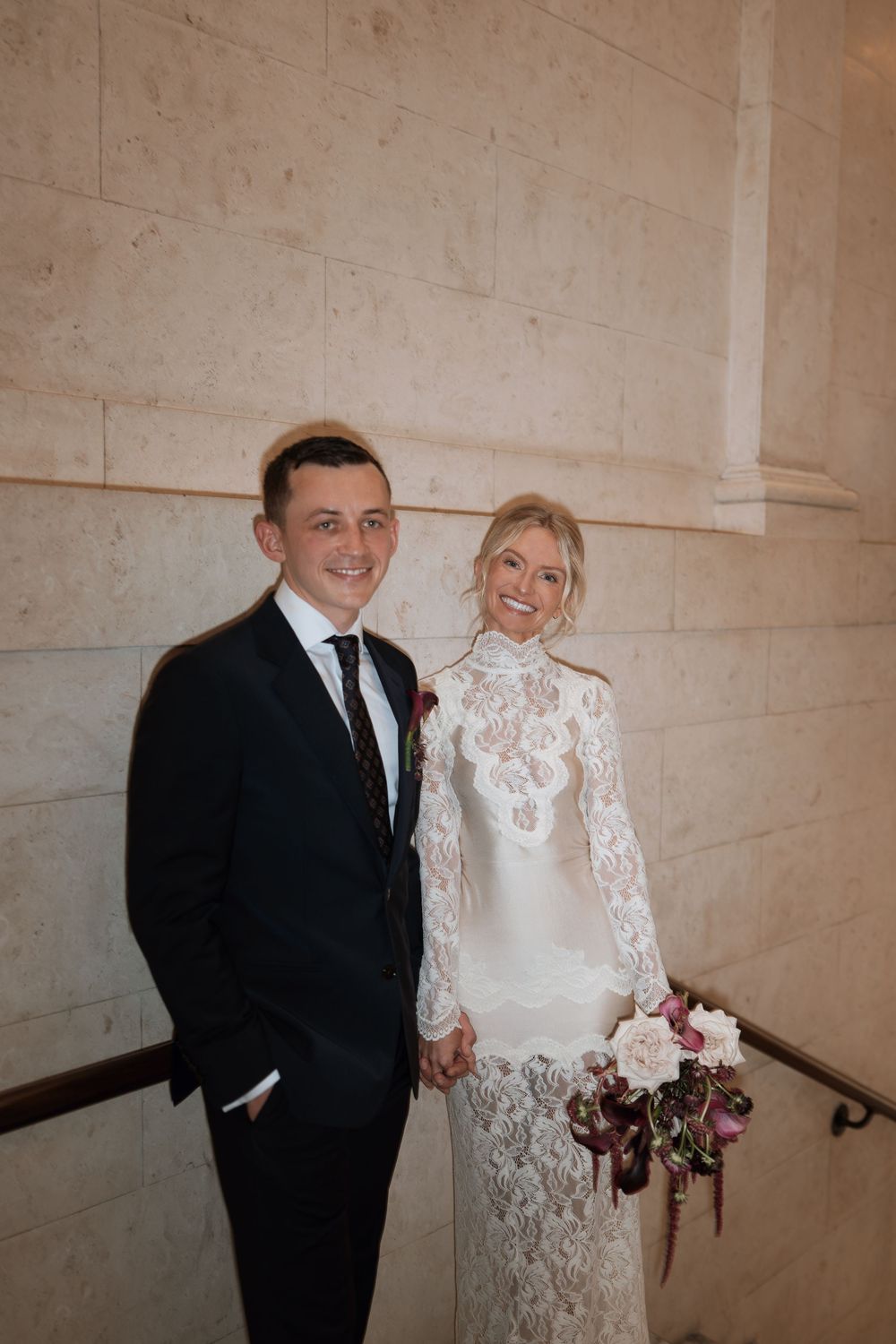 Couple posing on stairs in wedding attire, groom in dark suit, bride in lace long-sleeve white gown holding bouquet.