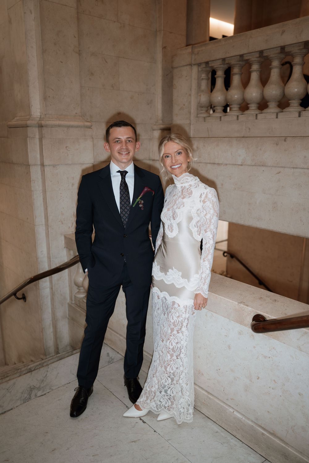 Couple posing on marble staircase, groom in navy suit, bride in white lace high-neck gown, elegant venue.