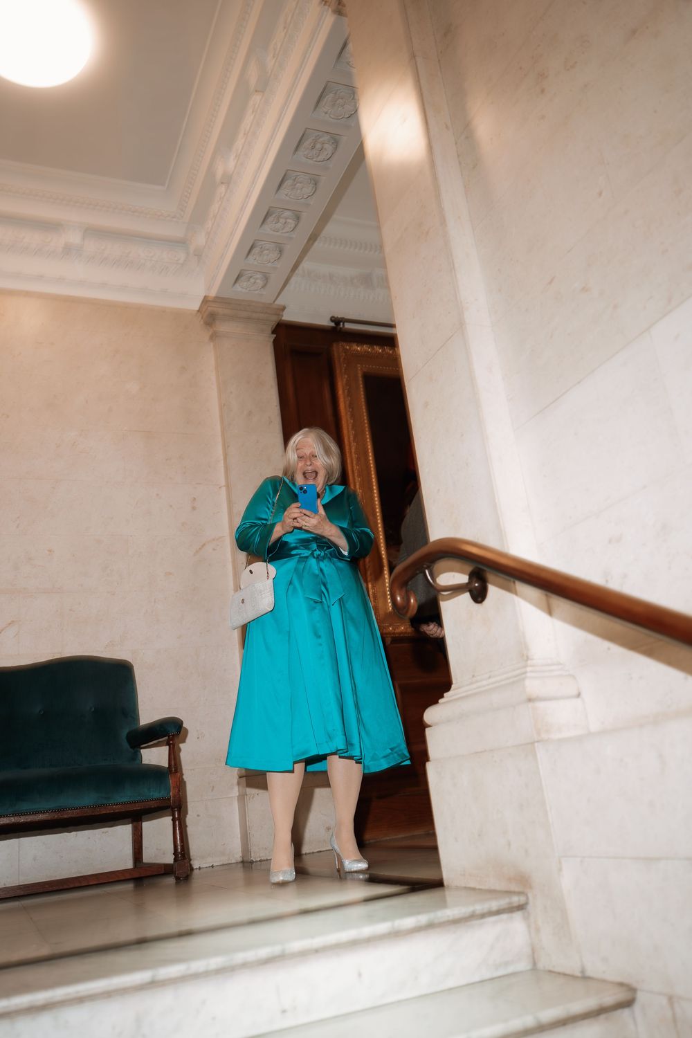 Elegant senior woman in teal dress descending marble stairs in a grand building, carrying a white purse.