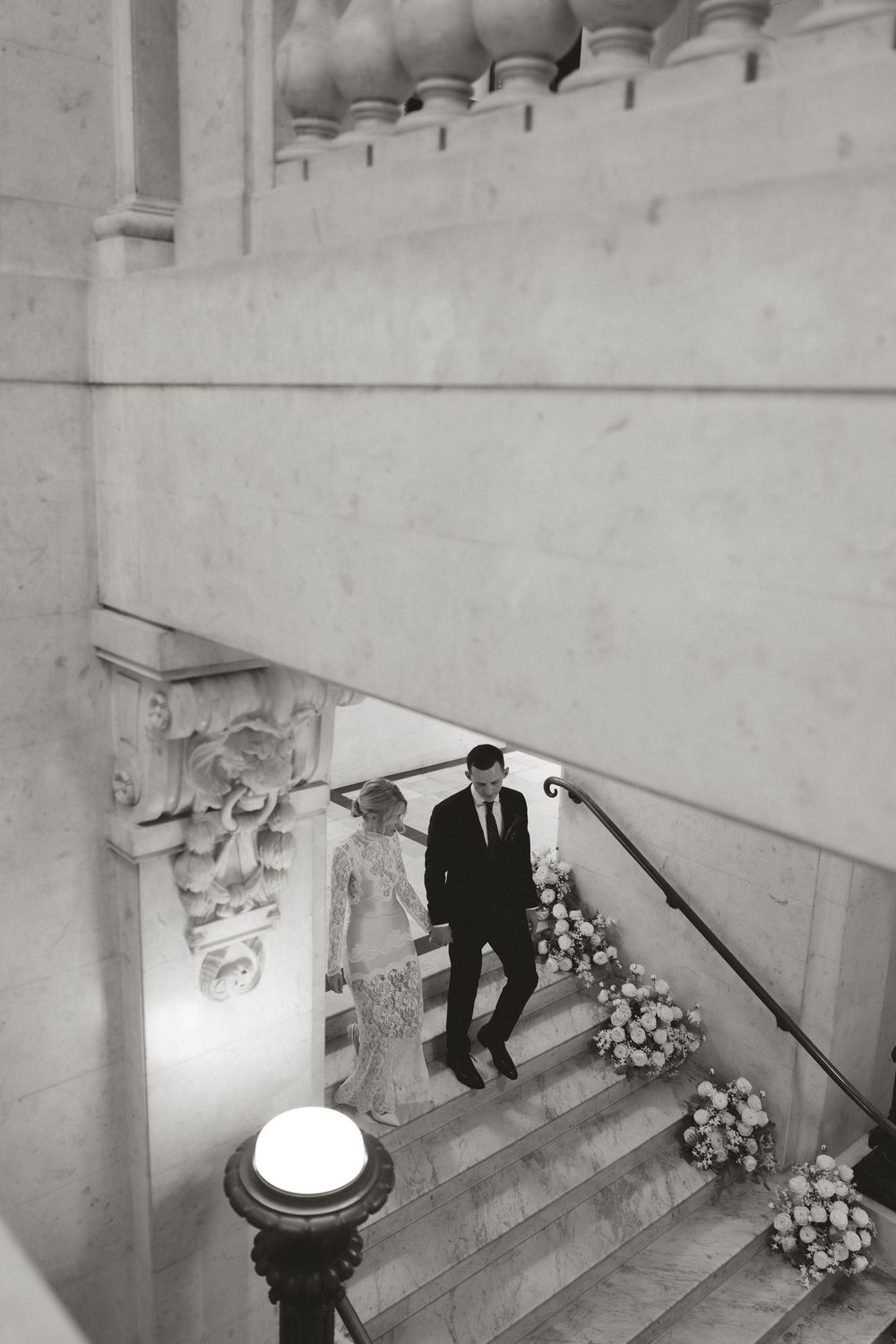 Black and white photo of a couple in wedding attire descending an elegant marble staircase adorned with floral arrangements.
