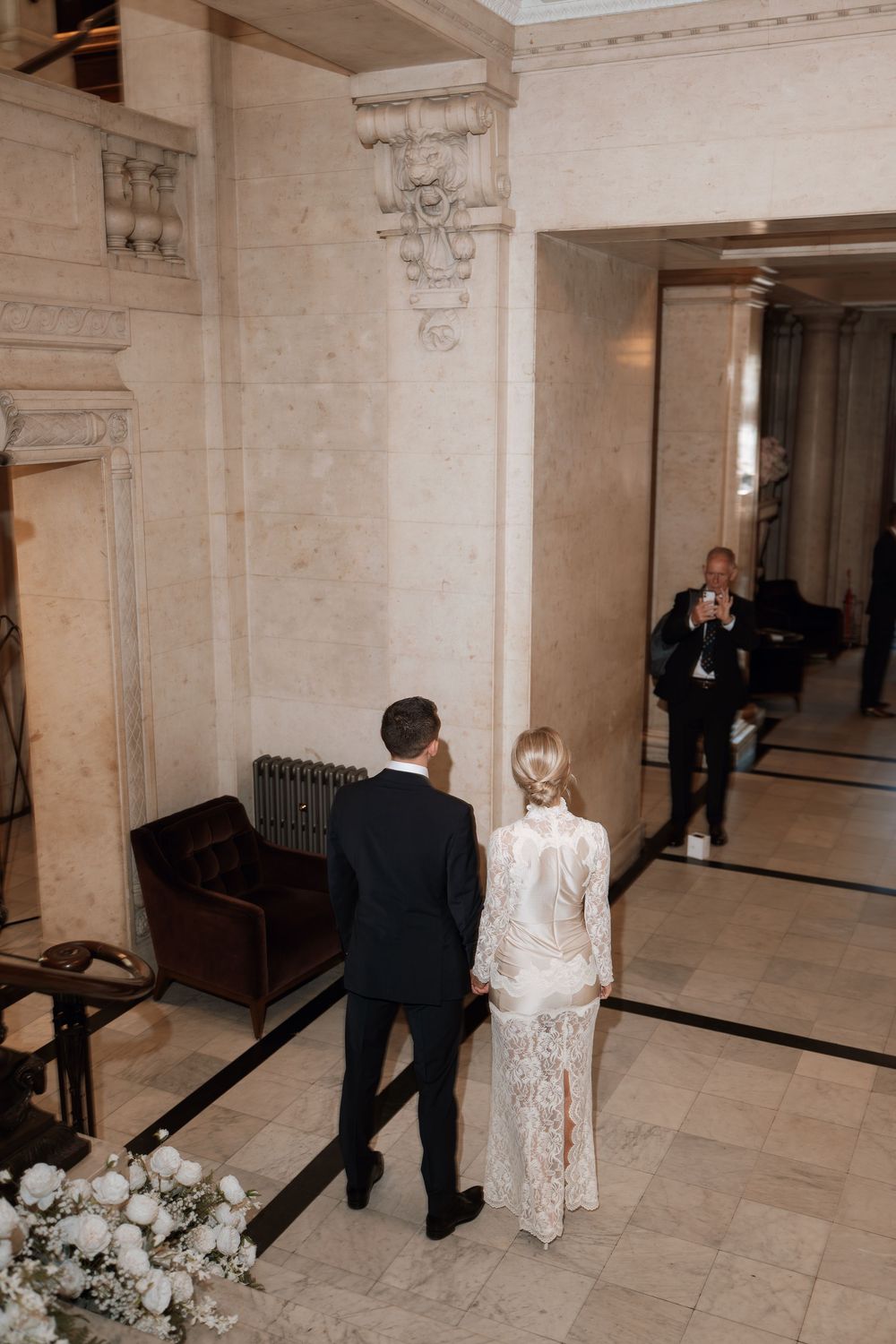 Bride and groom walking hand in hand through elegant marble hall, photographer capturing their wedding day moment.