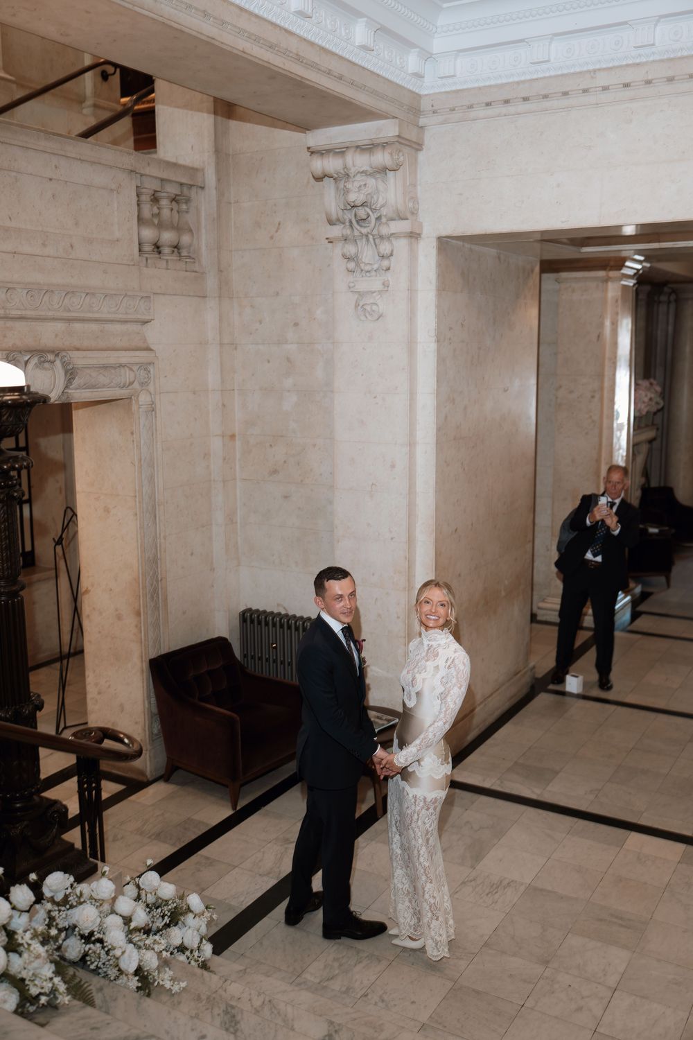 Bride and groom hold hands in elegant marble hall, she wears lace gown, he wears dark suit, guest photographs them.