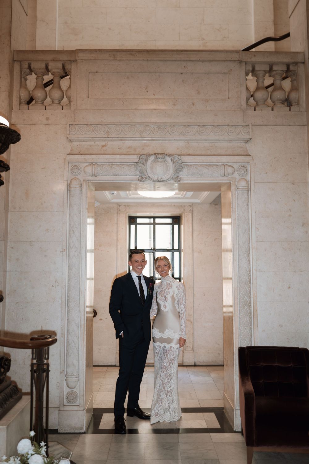 Bride and groom pose elegantly in white lace gown and dark suit inside a grand marble building.