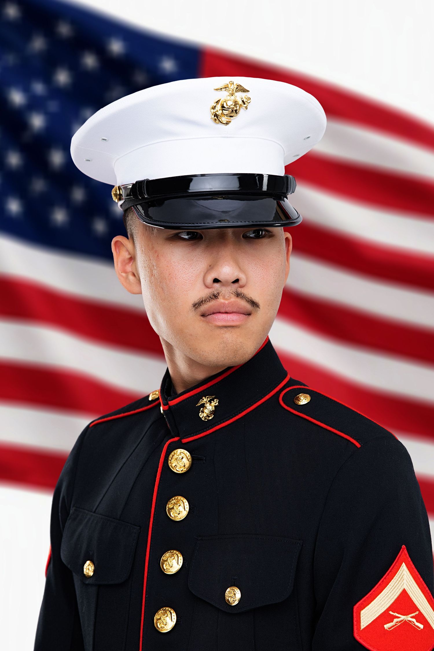 A young US Marine in dress blues uniform stands at attention before an American flag.