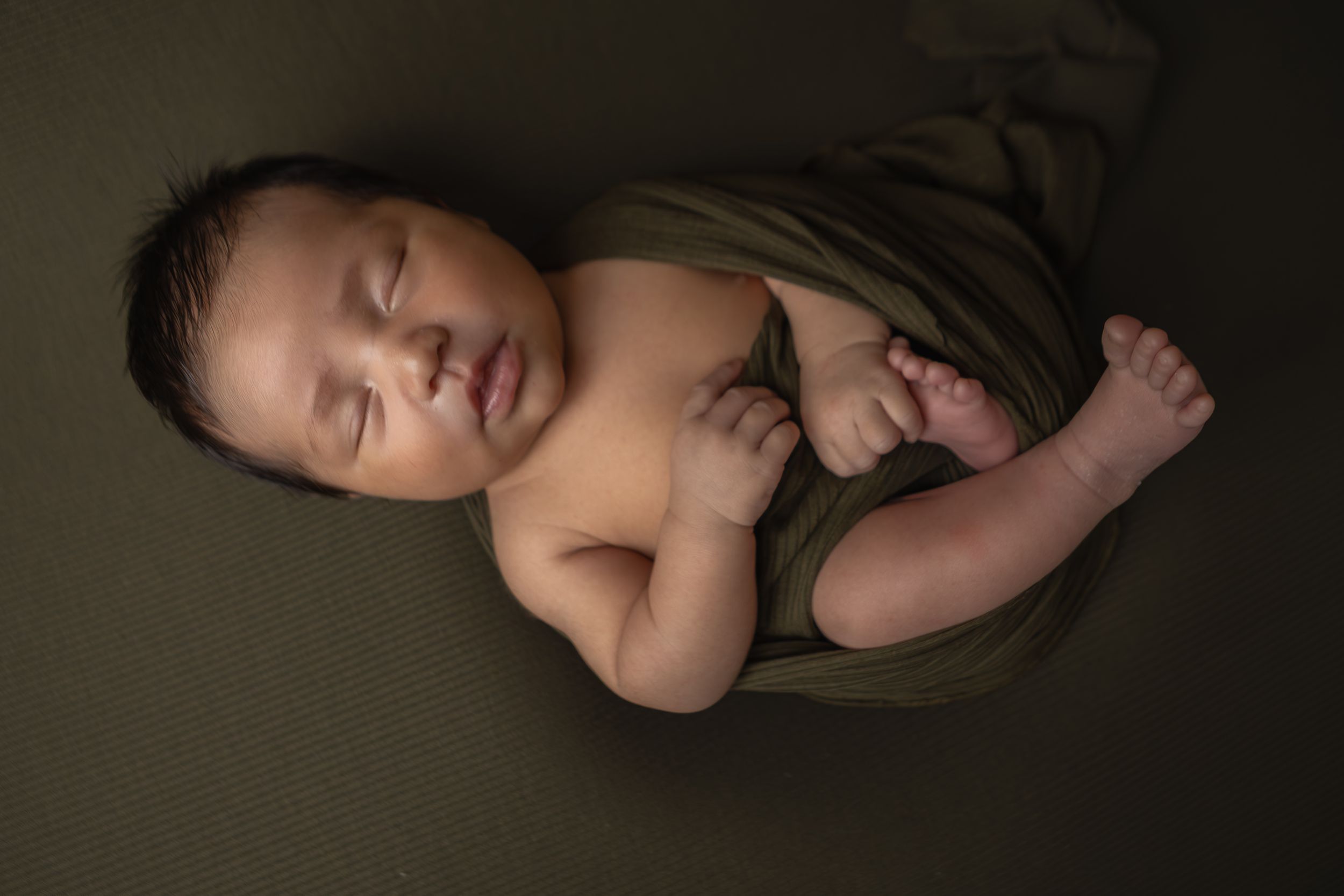 Serene newborn baby boy swaddled in olive green wrap sleeping on matching dark green backdrop.