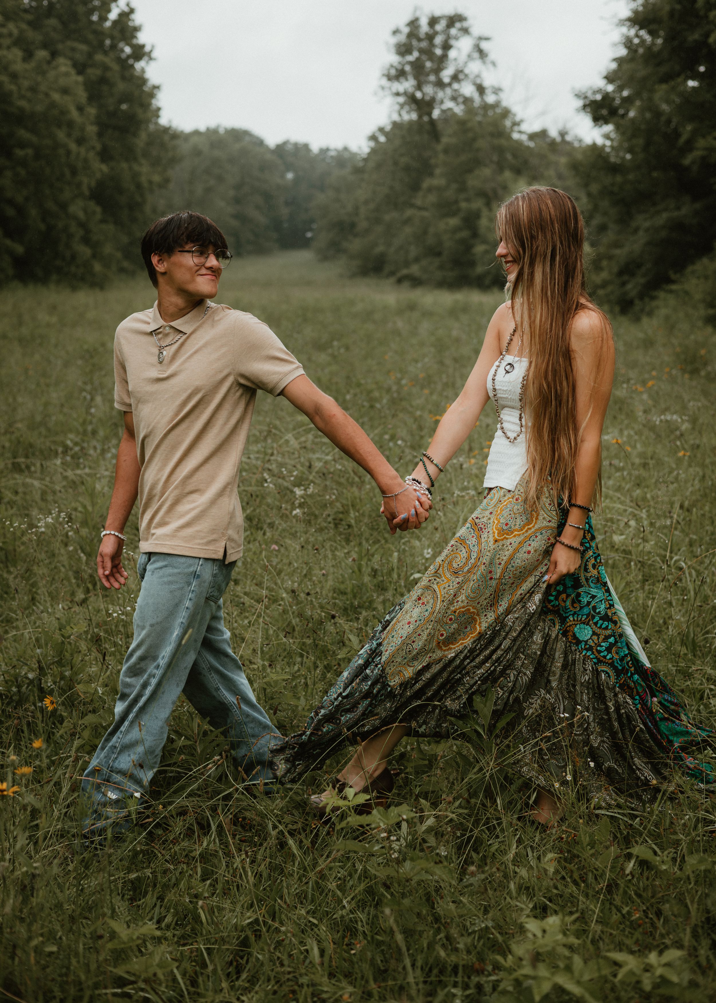 A young couple holds hands while walking through a lush green meadow, smiling at each other.