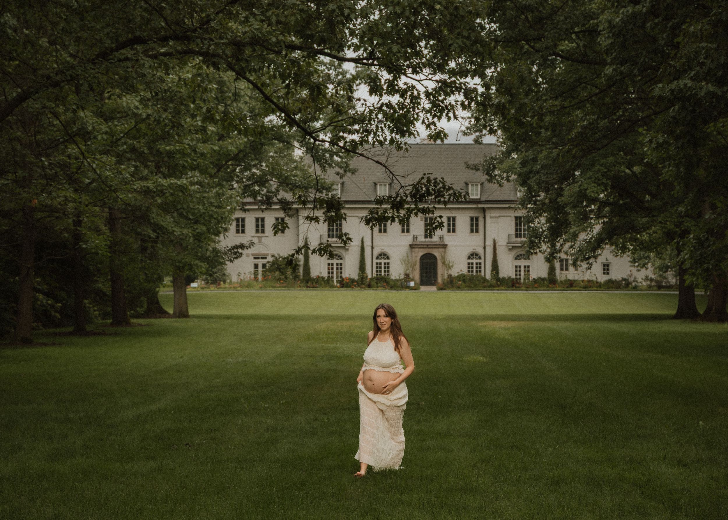 A pregnant woman in a white lace gown stands on a lush green lawn in front of an elegant estate.